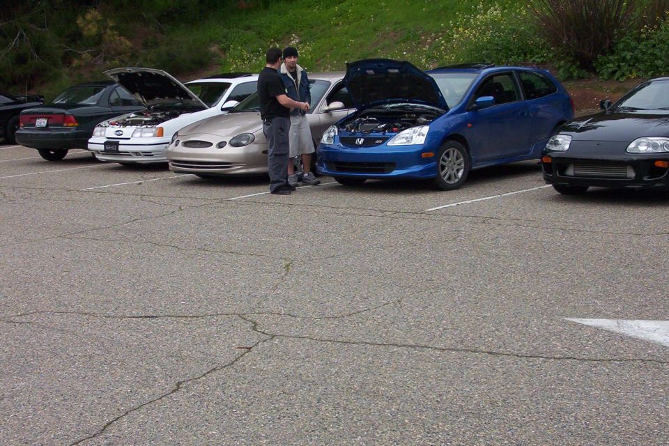 Several cars parked in a lot, including a black sports car, a blue hatchback, and a tan sedan. Two people stand by the blue car
