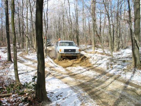 1995 Ford Bronco - A white pickup truck navigates a muddy, snow-dusted path through a forest, with another vehicle following behind.