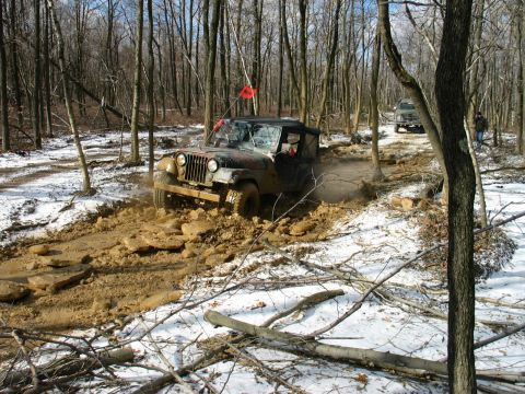 1995 Ford Bronco - A Jeep Wrangler struggles through a muddy section of a wooded trail in the snow.