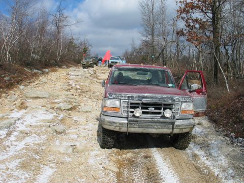 1995 Ford Bronco - A red Ford F-Series truck navigates a challenging off-road trail with other vehicles in the background.