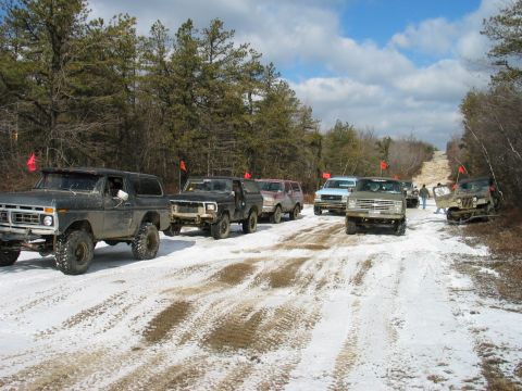 1995 Ford Bronco - A convoy of off-road vehicles navigates a snowy path through a wooded area under a partly cloudy sky.