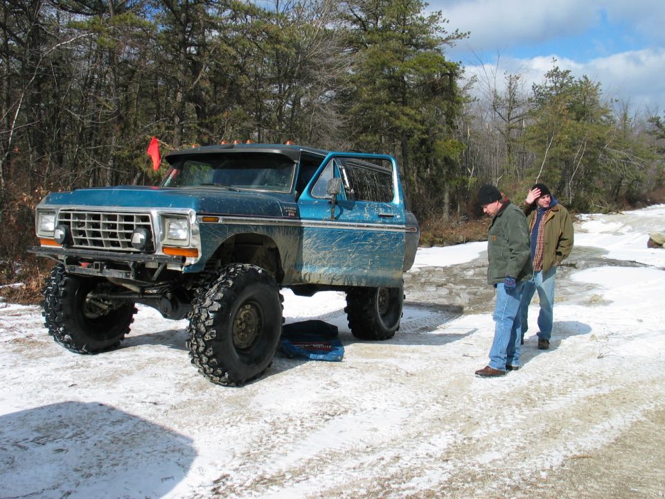 Blue Ford Bronco modified for off-road with large tires, two men standing nearby in a snowy wooded area