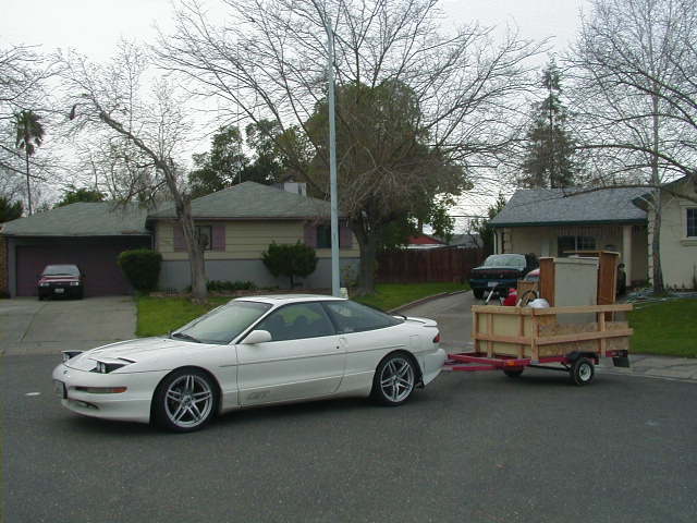 White Ford Probe coupe side view towing a small red trailer loaded with wood furniture on a suburban street