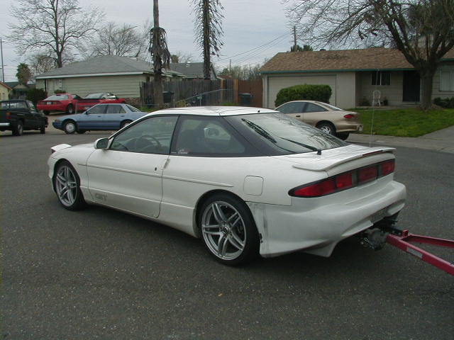 White Ford Probe coupe with aftermarket wheels and spoiler, rear 3/4 view, hooked to a red tow bar on a street