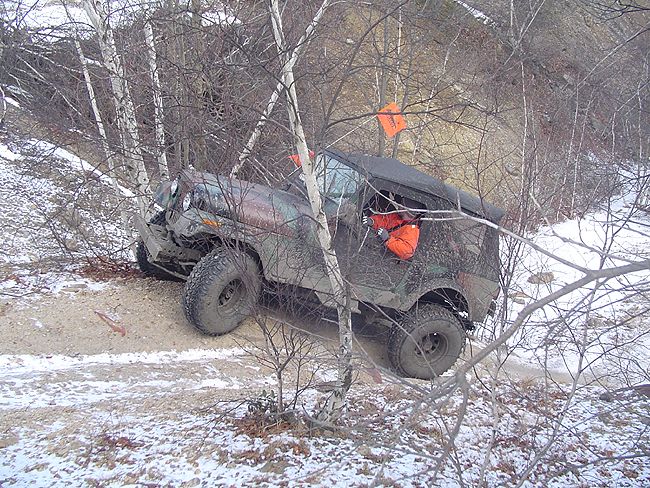 Off-road vehicle with large tires stuck between trees on a snowy, wooded trail. Driver wearing an orange jacket is visible inside