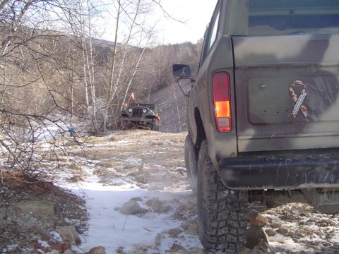 1979 Ford Bronco - Two off-road vehicles navigate a snowy, wooded path during daylight.