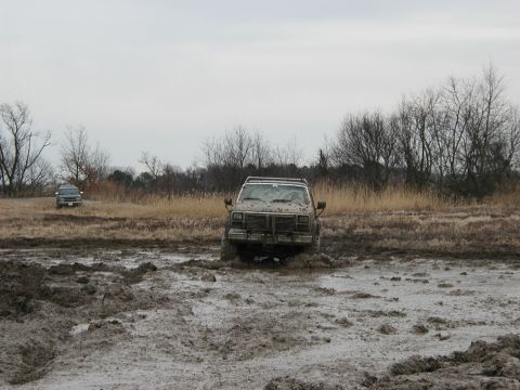 1985 Ford Bronco - Two off-road vehicles navigate a muddy terrain under an overcast sky.