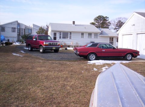 1995 Ford Bronco - Two vehicles, a red SUV and a red coupe, are parked in a residential driveway on a winter day.