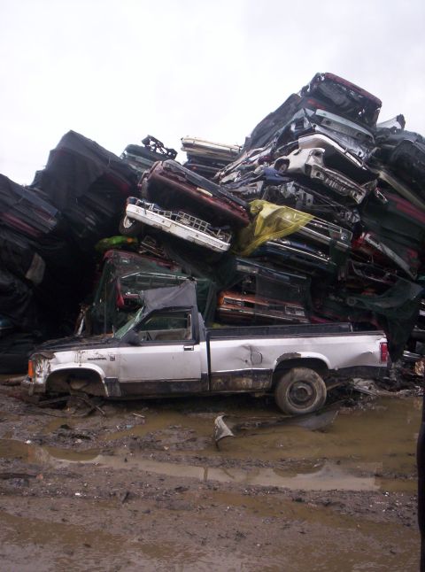 1995 Ford Bronco - A severely damaged pickup truck sits in a muddy junkyard, surrounded by stacks of crushed vehicles.