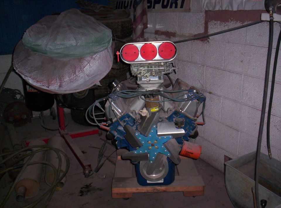 V8 engine with triple carb intake, blue fan, and orange oil filter. Engine is on a wooden stand