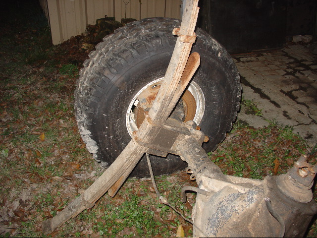 Off-road tire, leaf spring, and rusty axle/differential parts lying on leafy ground outdoors