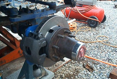 1986 Ford Bronco - Close-up of a vehicle's front wheel hub and brake assembly, with an air compressor in the background.