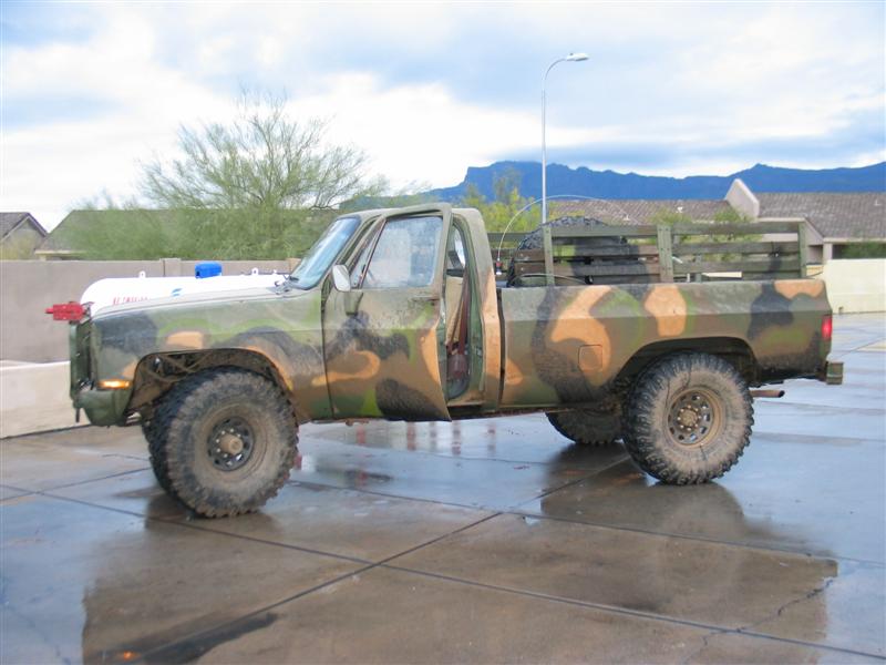 Camouflage-painted lifted Chevrolet pickup on mud tires parked on wet pavement with mountains behind