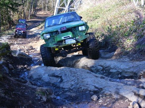1990 Ford Bronco - Multiple off-road vehicles tackle a challenging rocky terrain on a wooded trail.