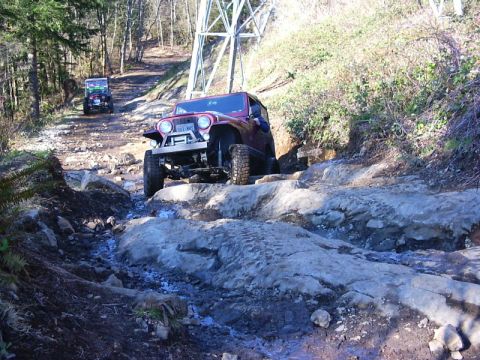 1990 Ford Bronco - Two Jeeps navigate a rocky off-road trail with steep inclines and dense foliage.