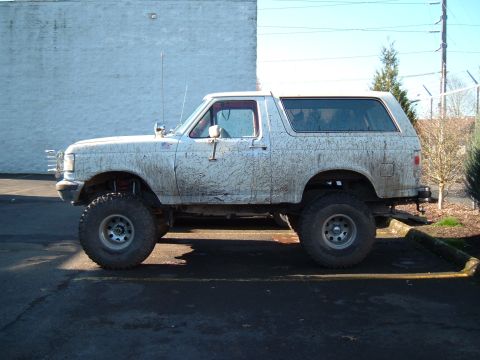 1989 Ford Bronco - Mud-covered, lifted Ford Bronco parked on asphalt. The vehicle's body style suggests a late 1980s model year.