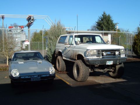 1989 Ford Bronco - A lifted Ford Bronco and a Triumph TR7 are parked side-by-side outdoors on a sunny day.