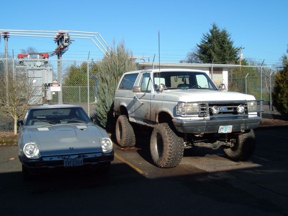 Lifted white Ford Bronco parked next to a silver Triumph TR7. Both vehicles are in a lot with a power substation in the background