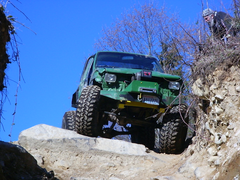 Green off-road SUV with large tires climbing over rocks, driver visible on right