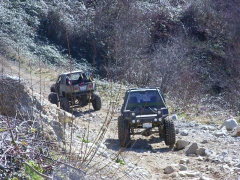 1990 Ford Bronco - Two heavily modified SUVs navigate a challenging, rocky off-road trail under a bright sky.