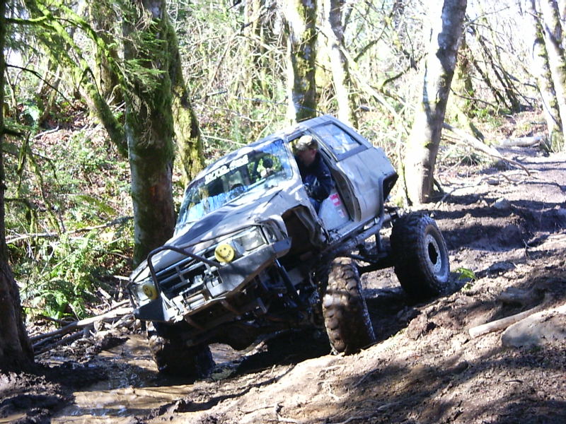 Modified SUV climbing a muddy trail in the woods. Driver visible inside