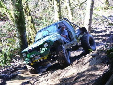 1990 Ford Bronco - Off-road vehicle navigating a muddy, wooded trail, with one wheel high off the ground.