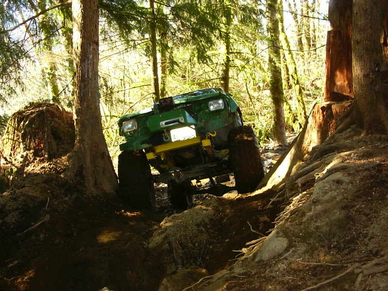 Green off-road vehicle navigating a steep, wooded trail with large rocks and trees