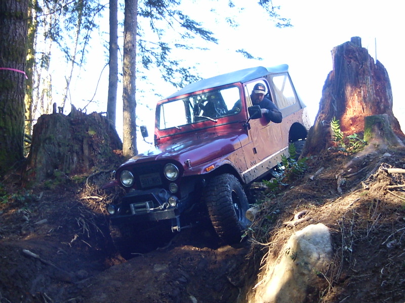 Red Jeep CJ-7 navigating a difficult off-road trail with large tree stumps and rocks