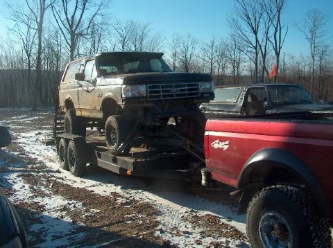 1995 Ford Bronco - A dark Ford Bronco is loaded onto a trailer, being pulled by a red pickup truck on a snowy day.