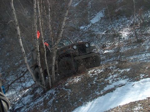 1995 Ford Bronco - An off-road vehicle is partially obscured by trees while climbing a muddy and snowy incline.