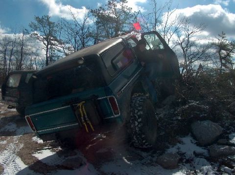 1995 Ford Bronco - A teal Ford Bronco is shown tilted on a snowy, rocky terrain, with a person entering the driver's side.