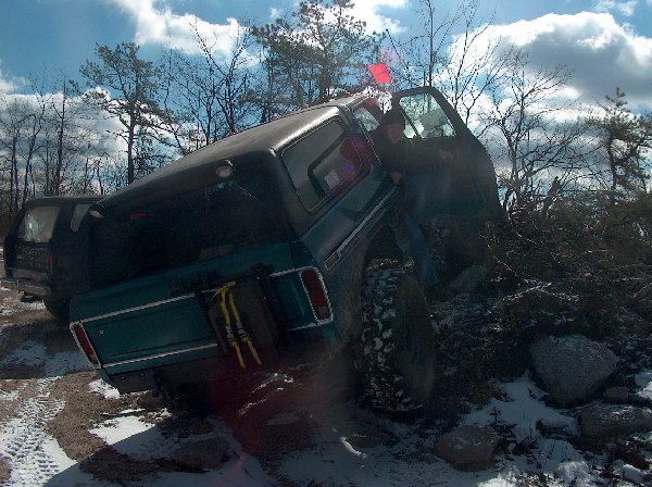Teal Ford Bronco with a black camper shell, tilted on a rocky incline with snow on the ground