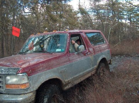 1995 Ford Bronco - A red and grey 1995 Ford Bronco navigates a muddy trail with a driver visible through the window.