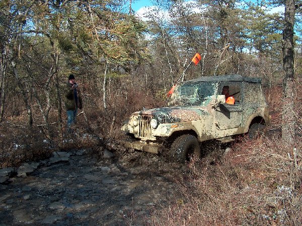Muddy Jeep CJ-7 driving through a wooded trail with a person standing in the background