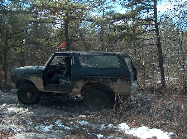 Mud-covered 1978 Ford Bronco with driver inside, parked on snowy, wooded trail