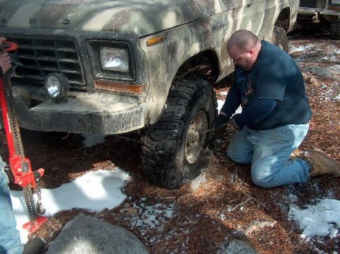 1995 Ford Bronco - A man works on a flat tire of a camouflage truck, with another vehicle visible in the background.