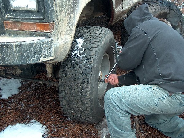 Person in hoodie and jeans working on a large tire with a lug wrench. Some snow on the ground