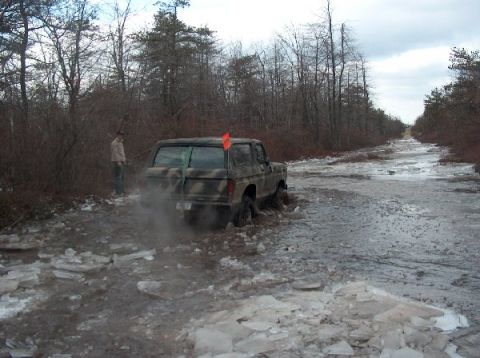 1995 Ford Bronco - A camouflaged SUV is stuck in icy mud on a dirt road surrounded by trees.