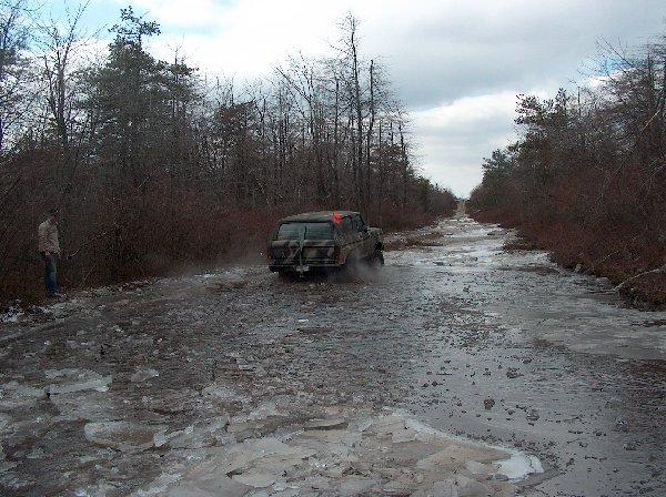 Camouflaged Ford Bronco driving on an icy, muddy road through a wooded area