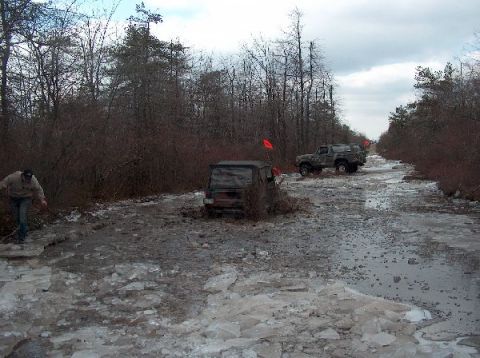 1995 Ford Bronco - Off-road vehicles tackle a challenging muddy and icy trail surrounded by bare trees under an overcast sky.