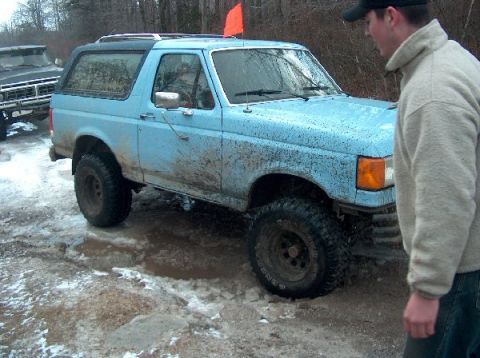 1995 Ford Bronco - A blue 1995 Ford Bronco navigates a muddy and icy terrain, with a person observing nearby.