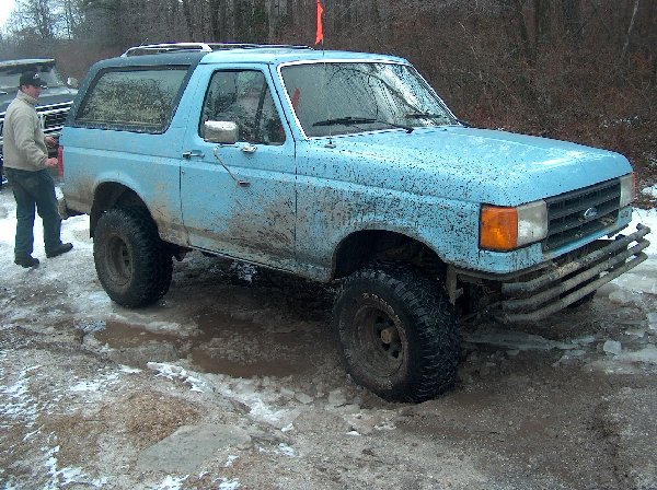 Mud-splattered blue 1987 Ford Bronco with aftermarket bumper, off-road tires, and a roof rack