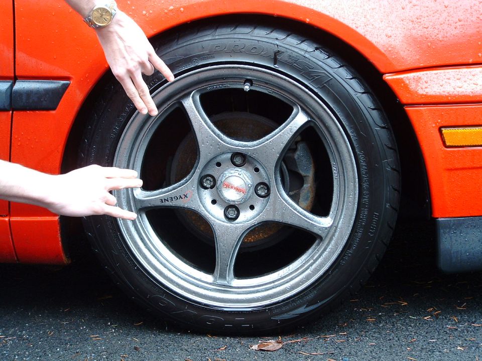 Close-up of a silver 5-spoke aftermarket wheel with 'XYGANE' visible on one spoke, mounted on a car with orange paint and black trim