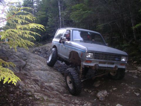 1984 Ford Bronco II - A silver 1984 Ford Bronco II navigates a challenging rocky incline on a wooded trail.