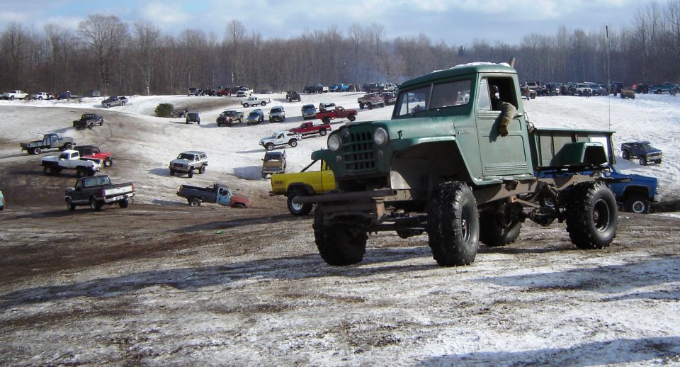 Green custom truck with big tires, parked in a snowy lot with other vehicles