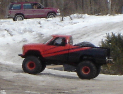 1978 Chevrolet C/K Pickup, lifted with large off-road tires, red and black paint scheme, driving in snow. Another SUV is visible in the background