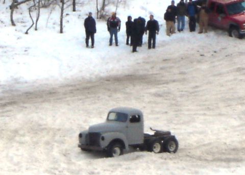 1978 Chevrolet C/K Pickup - A group of people watch a model truck in a snowy outdoor setting. A red pickup truck is visible in the background.