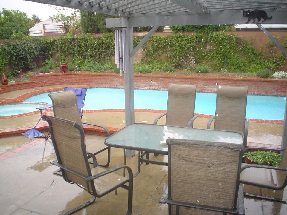 Backyard patio with a swimming pool and patio furniture under a pergola during a rain shower