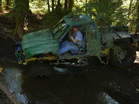 1990 Ford Bronco - A green off-road vehicle is stuck deep in mud and foliage in a wooded area.