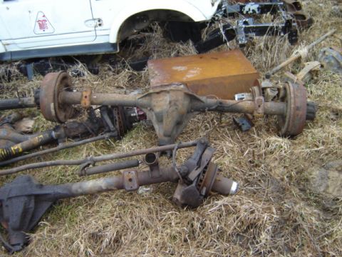 1992 Ford Bronco - Various automotive axles and suspension components are scattered on the ground amidst dry grass and debris.